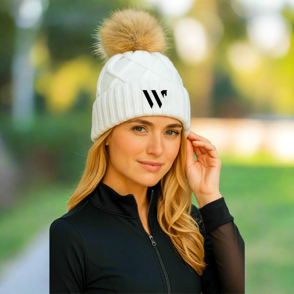 Woman wearing a white knit beanie with a pom-pom and a logo, outdoors.
