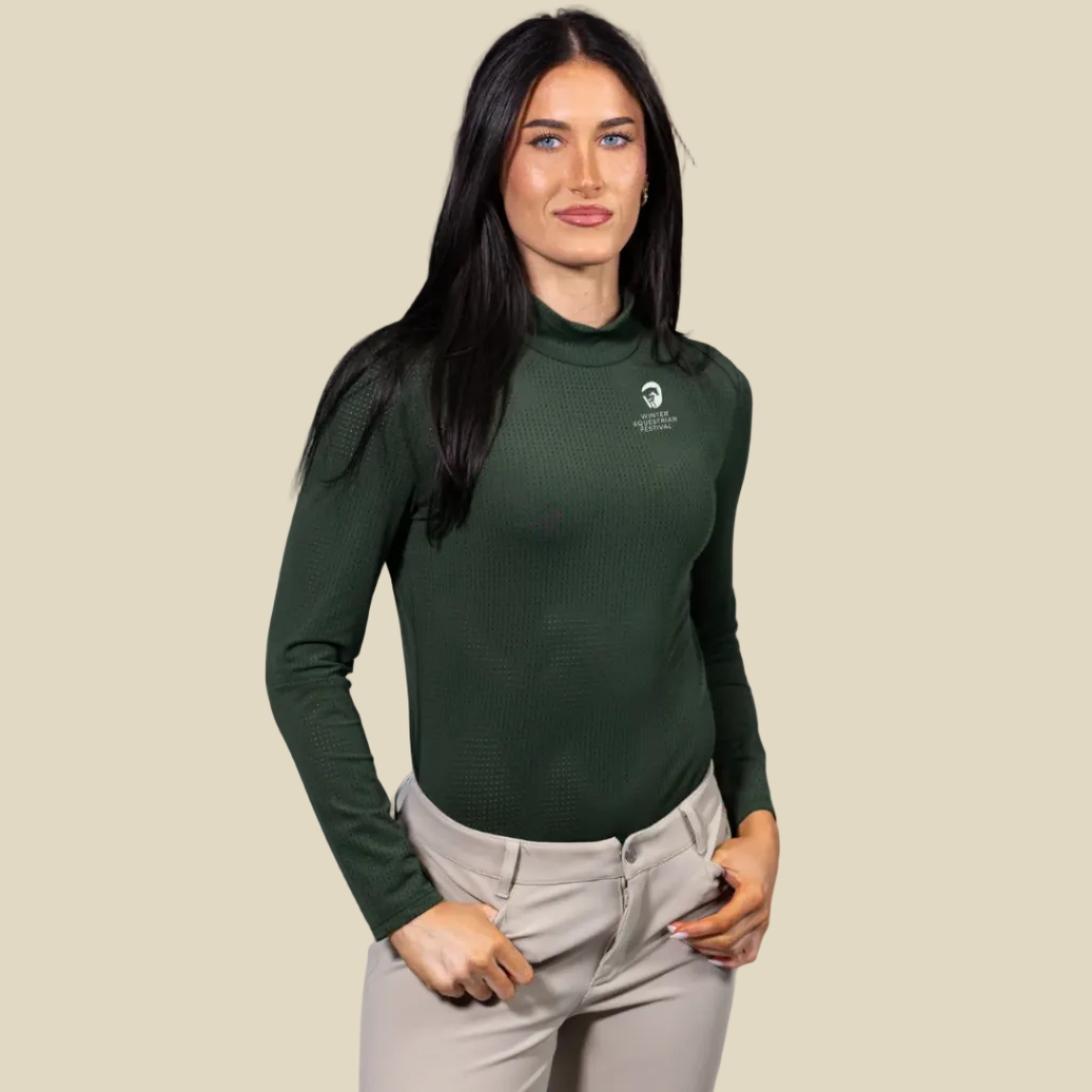 A woman modeling a dark green long-sleeve waffle-knit mock neck shirt with a small white logo on the left chest, paired with beige pants, against a plain tan background.