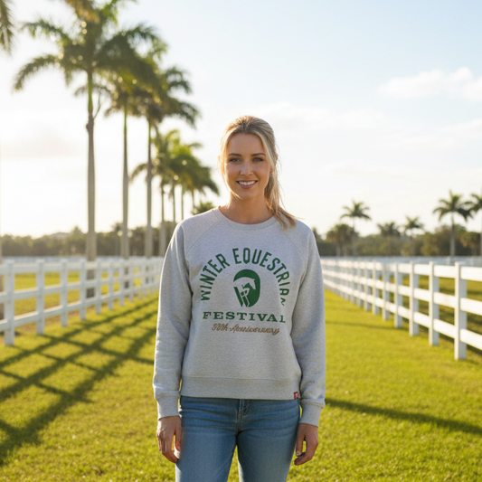 A woman wearing a light grey long-sleeve crewneck sweatshirt with a green and gold graphic on the chest reading 'Winter Equestrian Festival 50th Anniversary' on a horse farm